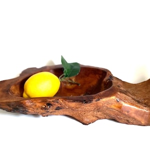 Puede incluir: Un cuenco de madera hecho a mano con una forma natural y orgánica. Contiene un limón amarillo brillante y una hoja verde. La madera tiene un color marrón cálido y veta visible.