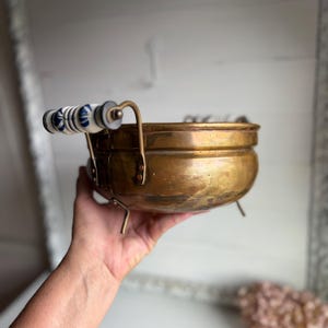 May include: A round brass bowl featuring a polished metallic finish, a wide rim, and three small metal feet. The bowl includes two side handles, each accented with white ceramic segments decorated with a blue floral pattern.