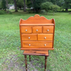 May include: A wooden tabletop cabinet with a scalloped top edge and six drawers featuring small white knobs. The bottom drawer is printed with the text: pain rustique, pain de mie, brioche, pain chocolat, choux à la crème, tarte aux amandes.