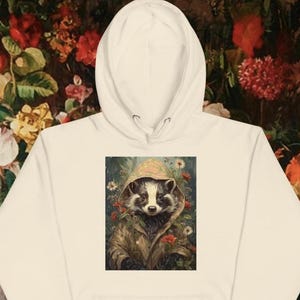 Puede incluir: Sudadera con capucha color crema con una ilustración de un mapache con impermeable, rodeado de flores. La ilustración está centrada en la parte delantera. El fondo es un estampado floral. La sudadera tiene capucha con cordón y bolsillo canguro.