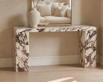 May include: A rectangular console table made of white marble with deep purple veining, featuring a tall arched mirror with a thin gold frame positioned above it, and a textured off-white ceramic vase holding thin branches.