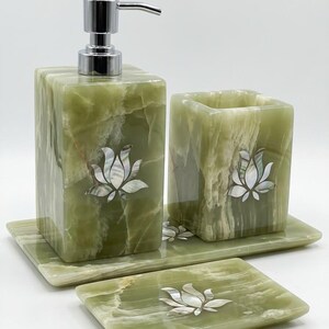 Può includere: Un set di accessori per il bagno realizzato in pietra verde chiaro con venature naturali. Il set comprende un dispenser di sapone rettangolare con pompa argentata, un portaspazzolino quadrato e due vassoi rettangolari, ciascuno decorato con un fiore di loto in madreperla intarsiato.
