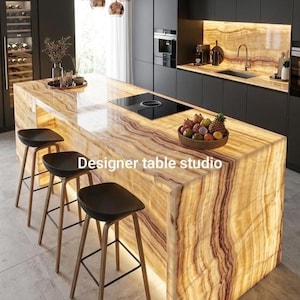 Może przedstawiać: Wyspa kuchenna z podświetlanym blatem z bursztynowego onyksu i bokami typu wodospad, z napisem Designer table studio. Trzy czarne hokery z drewnianymi nogami ustawione są przy wyspie, która zawiera czarną płytę grzewczą.