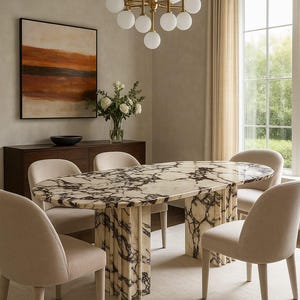 Op de afbeelding: Een ovale marmeren eettafel met een donker geaderd patroon, omringd door zes beige gestoffeerde stoelen. Boven hangt een gouden kroonluchter met bolvormige lampen. Een schilderij met oranje en bruine tinten hangt aan de muur.