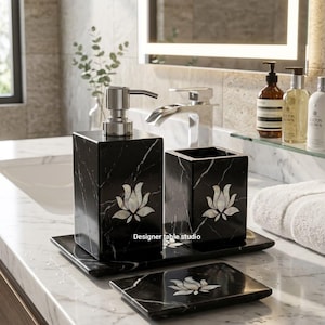 Può includere: Un set di accessori da bagno in marmo nero composto da un dispenser di sapone con pompa argentata, un portaspazzolino e un portasapone. Ogni pezzo è decorato con un intarsio floreale bianco e il testo Designer table studio è visibile.