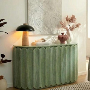 May include: A sage green semi-oval console table with a vertical fluted design. On top sits a black mushroom-shaped lamp with a cork base, a stack of books, a small terracotta-coloured vase, and a white vase containing dried pink branches.