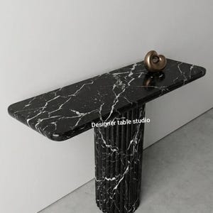 Puede incluir: Una mesa consola de mármol negro con vetas blancas, que presenta una superficie rectangular con bordes redondeados y una base de pedestal cilíndrica y estriada. Una pequeña escultura abstracta lisa de color bronce descansa sobre la superficie. El texto dice Designer table studio.