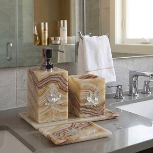 Può includere: Un set di accessori da bagno realizzato in pietra lucida con venature marroni e crema. Il set include un dispenser di sapone rettangolare con pompa argentata, un bicchiere quadrato e un portasapone rettangolare, decorati con un loto in madreperla.