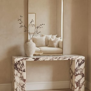 Pode incluir: Uma consola de mármore com um padrão bordô e branco. Um grande espelho arqueado com moldura dourada é montado acima da mesa. Um vaso bege com galhos está sobre a mesa, refletindo um sofá no espelho.
