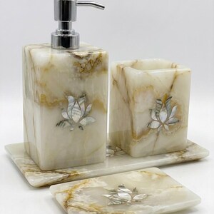 Può includere: Un set di accessori da bagno realizzato in pietra color crema con venature marroni, composto da un dispenser di sapone con pompa cromata, un portaspazzolino e un portasapone. Ogni pezzo è intarsiato con un fiore di loto in madreperla iridescente.