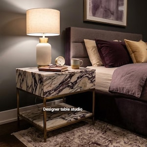 May include: A square bedside table featuring a white marble top and lower shelf with dark grey veining, supported by a thin gold metal frame. The table includes a single drawer and displays the text Designer table studio on the front.
