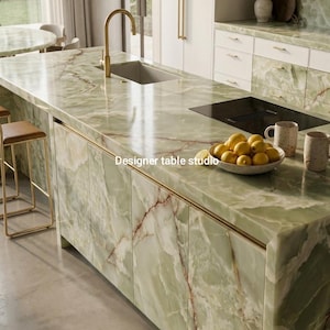 May include: A kitchen island featuring a polished green onyx countertop and cabinetry with prominent white and brown veining. The island includes a gold faucet, a sink, a black cooktop, and a bowl of lemons. Text reads Designer table studio.