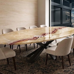 Puede incluir: Una mesa de comedor ovalada con un tablero de piedra ónix pulida en tonos amarillo claro y naranja, con una veta roja prominente en el centro. La mesa tiene una base de metal negro y está rodeada por ocho sillas tapizadas en beige.