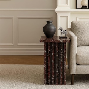 May include: A square side table with a red and brown marble top and base. A dark gray vase and a zebra figurine sit on top of the table. The table is next to a beige sofa.