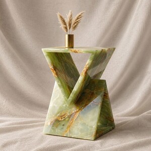 May include: A geometric side table crafted from polished green onyx with natural brown veining, featuring a triangular hourglass silhouette. A small, cylindrical metallic gold vase containing three dried pampas grass stems sits on top.
