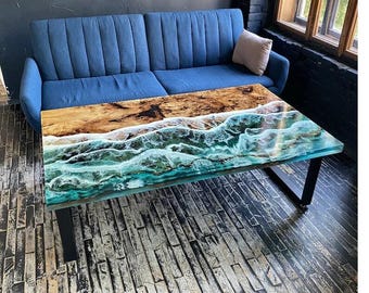 May include: A rectangular coffee table with a unique design. The top features a wood grain pattern transitioning into a turquoise and white wave design, encased in clear resin. The table has black metal legs and is placed in front of a blue sofa.