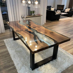 Puede incluir: Una mesa rectangular de madera con un diseño de río de resina epoxi transparente. La mesa tiene vetas de madera oscura y patas de metal negro. Está colocada sobre una alfombra blanca y esponjosa en una habitación bien iluminada.