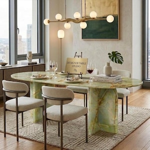 Könnte beinhalten: Ein ovaler Esstisch mit einer Tischplatte und einem Sockel aus grünem Onyx, umgeben von vier gepolsterten Stühlen mit dunklen Metallrahmen. Ein goldenes Schild auf dem Tisch trägt die Aufschrift DESIGNER TABLE STUDIO. Eine moderne Leuchte.
