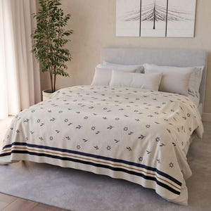 May include: A beige bedspread with a repeating pattern of dark blue leaves and stars. The bedspread has a border with navy blue and brown stripes. The bed is made with white pillows and a gray headboard. A potted plant and artwork are in the background.