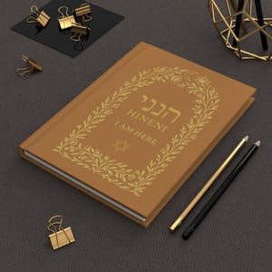 May include: A brown journal with gold lettering and a floral design. The text "HINENI I AM HERE" is displayed on the cover. Gold binder clips and pencils are also visible.