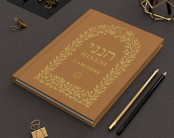 May include: A brown journal with gold lettering and a floral design. The text "HINENI I AM HERE" is displayed on the cover. Gold binder clips and pencils are also visible.