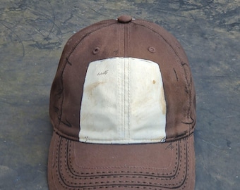 May include: A brown baseball cap with a white patch on the front. The cap has a curved brim and a button on top. The stitching is visible around the brim and the patch. The cap appears to be made of a durable fabric.