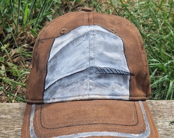 May include: Brown baseball cap with a light blue and gray design on the front. The brim has a light gray stripe. The cap is sitting on a wooden surface with green grass in the background.
