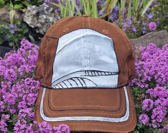 May include: A brown baseball cap with a white and gray design on the front. The cap has a curved brim and a silver trim. The cap is surrounded by purple flowers and green foliage.