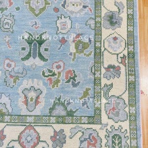 K&ouml;nnte beinhalten: Ausschnitt eines hellblauen Teppichs mit floralem Muster und cremefarbenem Rand. Der Teppich weist ein sich wiederholendes Design stilisierter Blumen in Gr&uuml;n-, Grau- und Rosat&ouml;nen auf. Der Text "HANDKNOTPLACE" ist auf dem Teppich zu sehen.