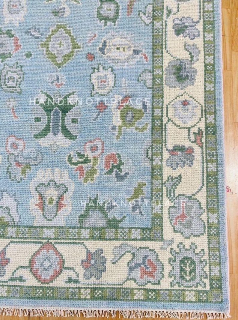 K&ouml;nnte beinhalten: Ausschnitt eines hellblauen Teppichs mit floralem Muster und cremefarbenem Rand. Der Teppich weist ein sich wiederholendes Design stilisierter Blumen in Gr&uuml;n-, Grau- und Rosat&ouml;nen auf. Der Text "HANDKNOTPLACE" ist auf dem Teppich zu sehen.