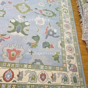 K&ouml;nnte beinhalten: Hellblauer Teppich mit cremefarbenem Rand und bunten Blumenmustern. Der Teppich hat ein Mittelfeld mit sich wiederholenden abstrakten Formen in Gr&uuml;n, Rot und Grau. Der Rand hat ein Blumendesign mit gr&uuml;nen und roten Akzenten. Der Text "HANDKNOTTPLACE" ist sichtbar.