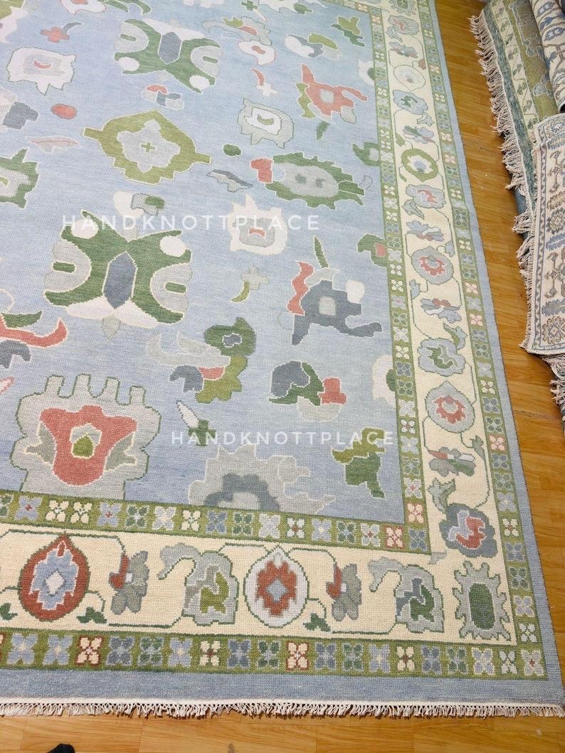 K&ouml;nnte beinhalten: Hellblauer Teppich mit cremefarbenem Rand und bunten Blumenmustern. Der Teppich hat ein Mittelfeld mit sich wiederholenden abstrakten Formen in Gr&uuml;n, Rot und Grau. Der Rand hat ein Blumendesign mit gr&uuml;nen und roten Akzenten. Der Text "HANDKNOTTPLACE" ist sichtbar.