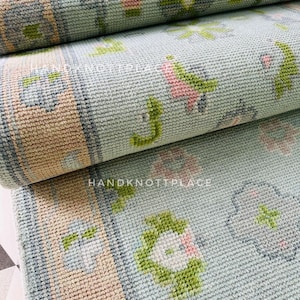 May include: Close-up of a rolled-up rug with a light blue background and floral patterns in pink, green, and white. The border is a tan color with a gray outline. The text "HANDKNOTTPLACE" is visible on the rug.