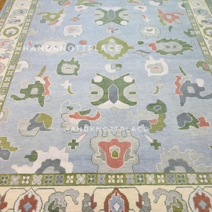 K&ouml;nnte beinhalten: Ein gro&szlig;er, rechteckiger Teppich mit hellblauem Feld und detailliertem Rand. Der Teppich weist ein sich wiederholendes Muster aus floralen und geometrischen Formen in Gr&uuml;n-, Rot-, Grau- und Cremet&ouml;nen auf. Der Text "HANDKNOTPLACE" ist sichtbar.