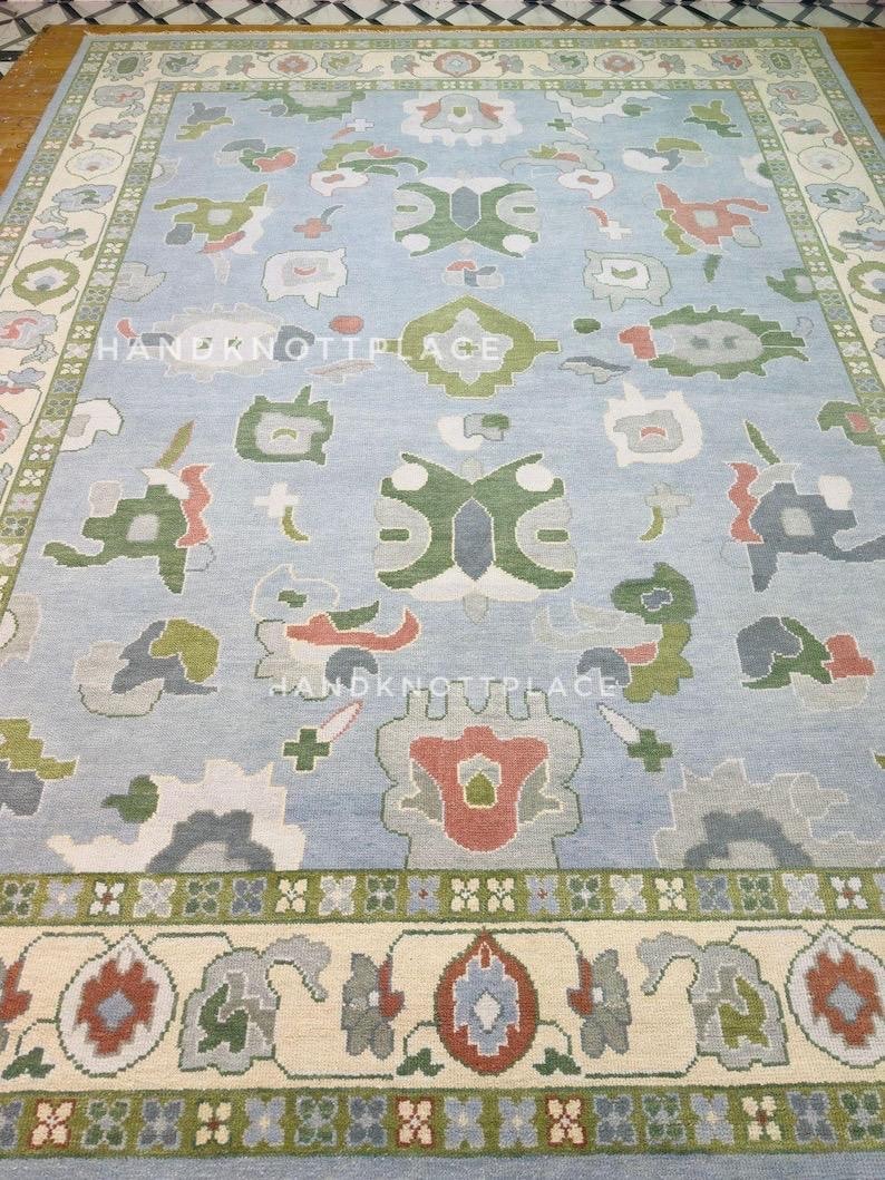 K&ouml;nnte beinhalten: Ein gro&szlig;er, rechteckiger Teppich mit hellblauem Feld und detailliertem Rand. Der Teppich weist ein sich wiederholendes Muster aus floralen und geometrischen Formen in Gr&uuml;n-, Rot-, Grau- und Cremet&ouml;nen auf. Der Text "HANDKNOTPLACE" ist sichtbar.