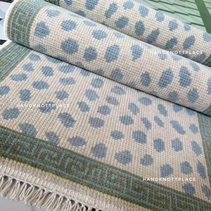 May include: Rolled-up area rugs with a cream background and blue polka dots. The rugs have a green border with a geometric pattern and fringe. The text "HANDKNOTTPLACE" is visible on the rug.