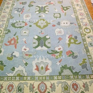 K&ouml;nnte beinhalten: Ein rechteckiger, hellblauer Teppich mit cremefarbenem Rand und komplizierten floralen und geometrischen Mustern in Gr&uuml;n-, Rot- und Graut&ouml;nen. Der Teppich tr&auml;gt den Text "HANDKNOTTPLACE".