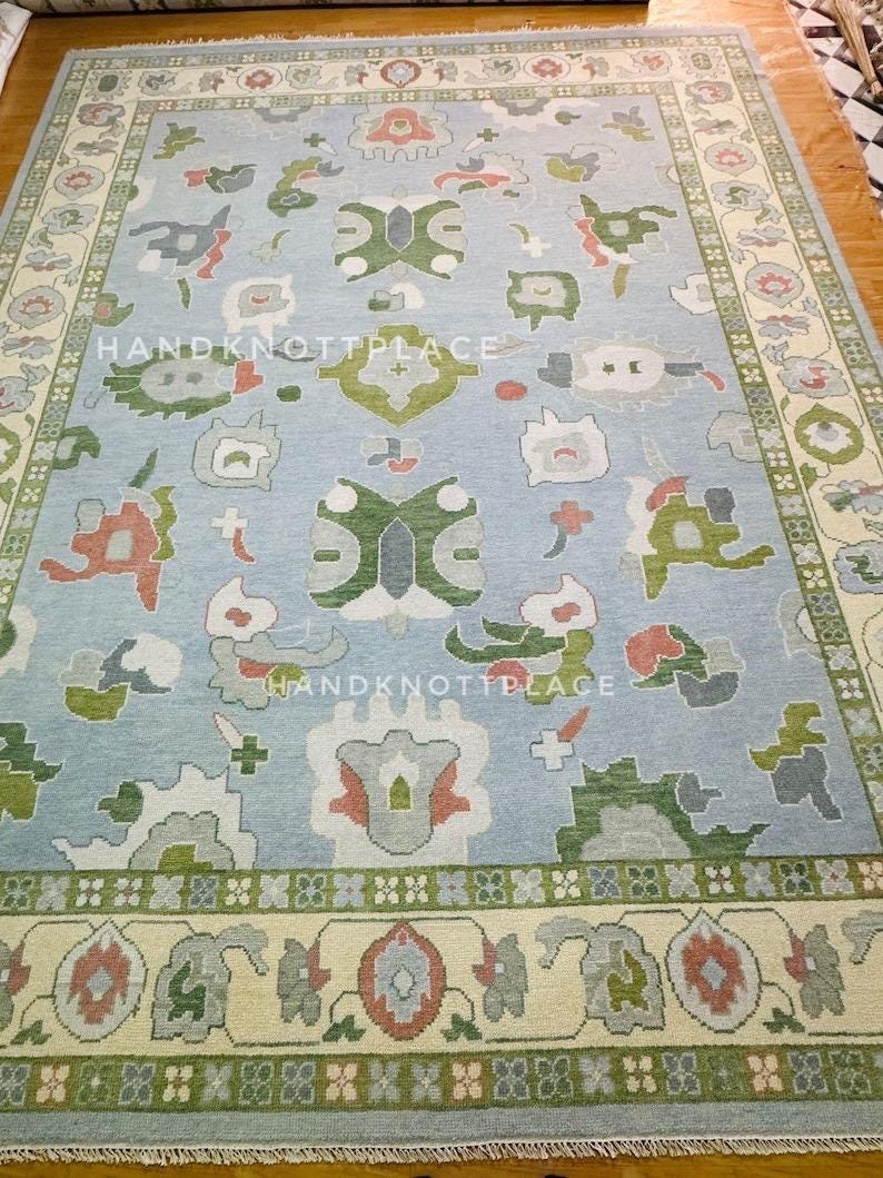 K&ouml;nnte beinhalten: Ein rechteckiger, hellblauer Teppich mit cremefarbenem Rand und komplizierten floralen und geometrischen Mustern in Gr&uuml;n-, Rot- und Graut&ouml;nen. Der Teppich tr&auml;gt den Text "HANDKNOTTPLACE".