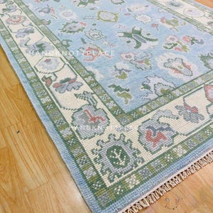 K&ouml;nnte beinhalten: Rechteckiger, hellblauer Teppich mit detailliertem Blumenmuster und cremefarbenem Rand. Der Rand ist mit gr&uuml;nen und grauen geometrischen Designs akzentuiert. Der Teppich hat an einem Ende einen Fransen und der Text "HAND NOT PLACE" ist sichtbar.
