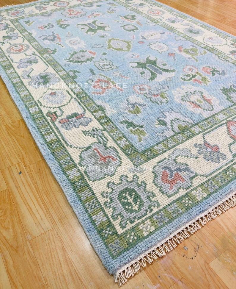 K&ouml;nnte beinhalten: Rechteckiger, hellblauer Teppich mit detailliertem Blumenmuster und cremefarbenem Rand. Der Rand ist mit gr&uuml;nen und grauen geometrischen Designs akzentuiert. Der Teppich hat an einem Ende einen Fransen und der Text "HAND NOT PLACE" ist sichtbar.