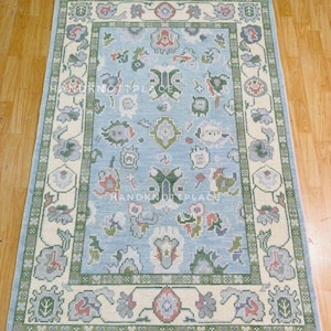 K&ouml;nnte beinhalten: Rechteckiger Teppich mit hellblauem Feld und cremefarbenem Rand. Der Teppich zeigt florale und geometrische Muster in Gr&uuml;n-, Rot- und Graut&ouml;nen. Der Text "HANDKNOT PLACE" ist auf dem Teppich zu sehen.