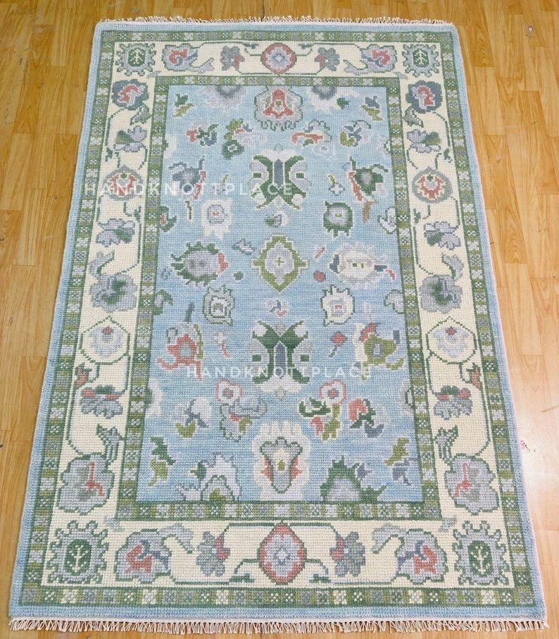 K&ouml;nnte beinhalten: Rechteckiger Teppich mit hellblauem Feld und cremefarbenem Rand. Der Teppich zeigt florale und geometrische Muster in Gr&uuml;n-, Rot- und Graut&ouml;nen. Der Text "HANDKNOT PLACE" ist auf dem Teppich zu sehen.