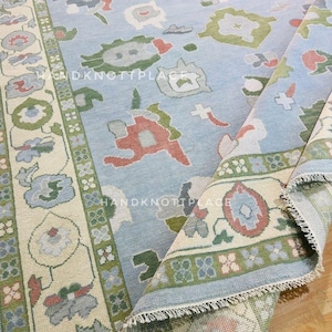 K&ouml;nnte beinhalten: Hellblauer, handgekn&uuml;pfter Teppich mit floralem und geometrischem Muster. Der Teppich hat einen cremefarbenen Rand mit gr&uuml;nen und roten Akzenten. Der Text "HANDKNOTTPLACE" und "DKNOTTPLACE" ist sichtbar.