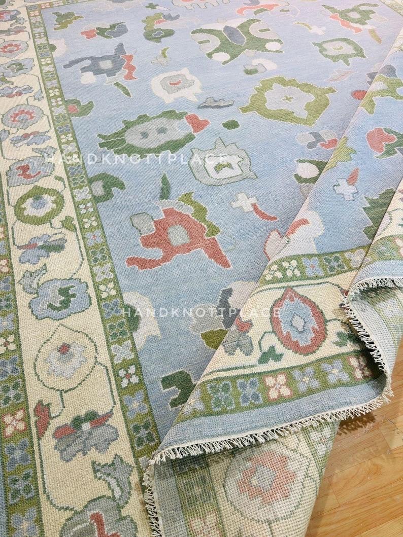 K&ouml;nnte beinhalten: Hellblauer, handgekn&uuml;pfter Teppich mit floralem und geometrischem Muster. Der Teppich hat einen cremefarbenen Rand mit gr&uuml;nen und roten Akzenten. Der Text "HANDKNOTTPLACE" und "DKNOTTPLACE" ist sichtbar.