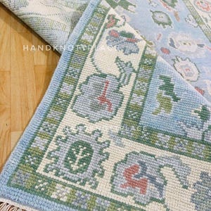 K&ouml;nnte beinhalten: Ein hellblau-cremefarbener Teppich mit floralem Muster und gr&uuml;nem Rand. Der Teppich ist teilweise aufgerollt und gibt die Unterseite frei. Der Text "HANDKNOTTED PLACE" ist auf dem Teppich zu sehen.