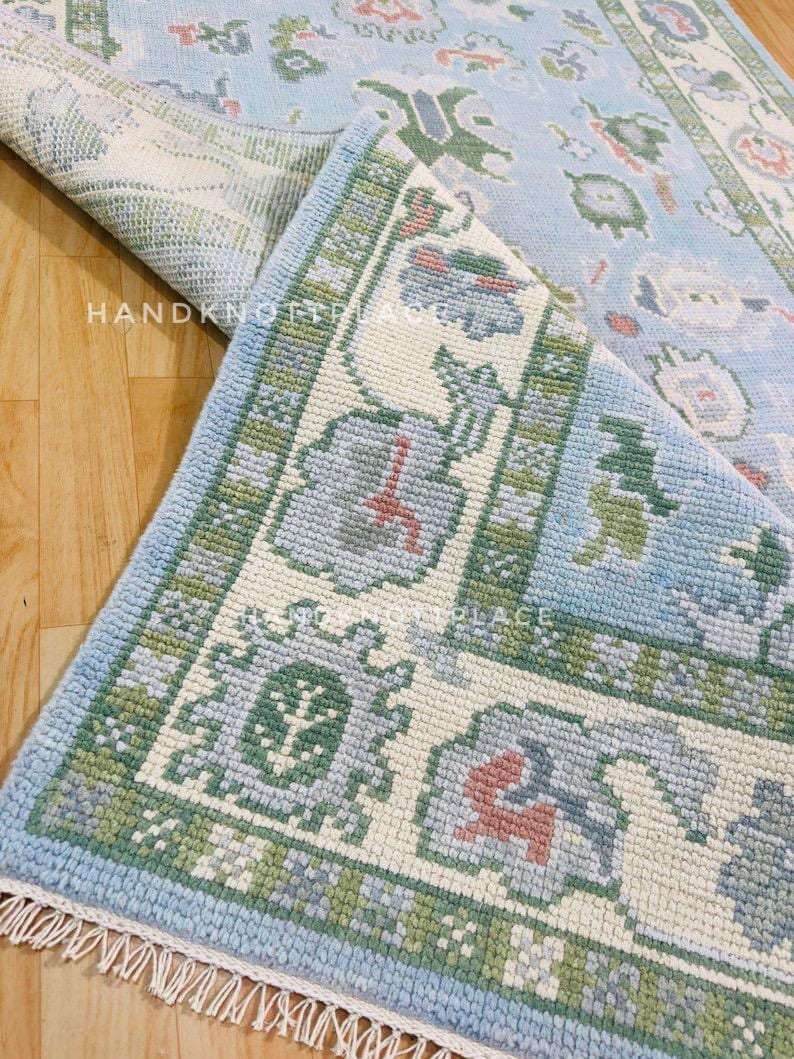 K&ouml;nnte beinhalten: Ein hellblau-cremefarbener Teppich mit floralem Muster und gr&uuml;nem Rand. Der Teppich ist teilweise aufgerollt und gibt die Unterseite frei. Der Text "HANDKNOTTED PLACE" ist auf dem Teppich zu sehen.