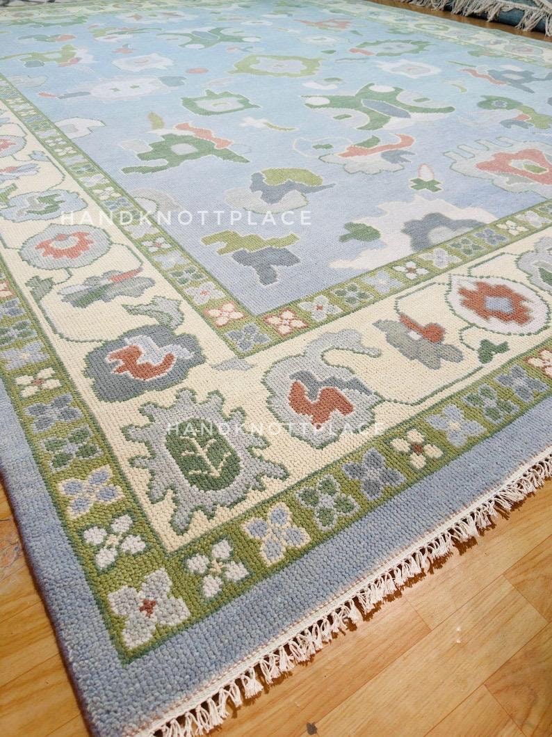 K&ouml;nnte beinhalten: Ein hellblauer Teppich mit cremefarbenem Rand und blauem Au&szlig;enrand. Der Teppich weist ein sich wiederholendes Muster aus floralen und abstrakten Formen in Gr&uuml;n-, Rot- und Graut&ouml;nen auf. Der Teppich hat einen Fransenrand und den Text "HANDKNOTTPLACE".