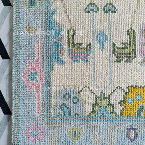 May include: Close-up of a hand-knotted rug with a cream-colored central field and a light blue border. The rug features intricate patterns in shades of blue, pink, yellow, and green. The design includes floral and geometric motifs, with a fringed edge.
