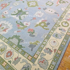 K&ouml;nnte beinhalten: Rechteckiger, hellblauer Teppich mit einem Blumenmuster in Gr&uuml;n-, Rot- und Graut&ouml;nen. Der Teppich hat einen cremefarbenen Rand mit einem sich wiederholenden Blumendesign und dem Text "HANDKNOTTPLACE".