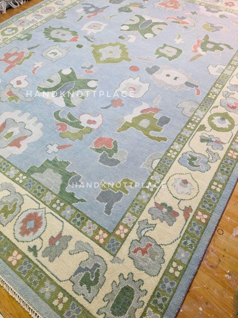 K&ouml;nnte beinhalten: Rechteckiger, hellblauer Teppich mit einem Blumenmuster in Gr&uuml;n-, Rot- und Graut&ouml;nen. Der Teppich hat einen cremefarbenen Rand mit einem sich wiederholenden Blumendesign und dem Text "HANDKNOTTPLACE".